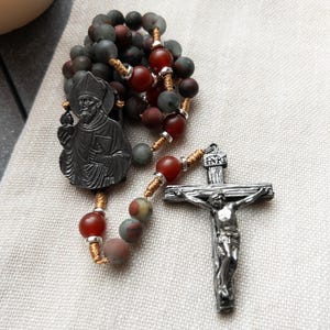 Op de afbeelding: Een rozenkrans met donkerrode, grijze en gespikkelde kralen. De rozenkrans heeft een metalen kruisbeeld en een metalen hanger met een religieuze figuur. De kralen zijn geregen op goudkleurig draad.
