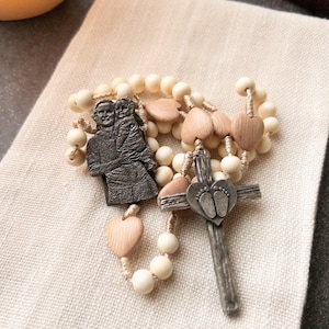 Puede incluir: Un rosario con cuentas de color crema, cuentas de madera en forma de corazón y un colgante de cruz plateado. La cruz presenta huellas de bebé. Un colgante oscuro representa una figura sosteniendo a un niño. El rosario está sobre una tela color crema.