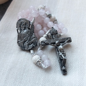 May include: A rosary with light pink rose quartz beads, silver-toned metal crucifix, and a saint medal. The rosary features decorative white beads and is laid on a light-colored fabric.