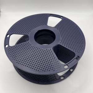 Könnte beinhalten: Eine dunkelblaue 3D-Drucker-Filamentspule. Die Spule hat ein perforiertes Design mit zahlreichen kleinen Löchern und einer zentralen kreisförmigen Öffnung. Die Spule besteht aus Kunststoff und ist zur Aufnahme von 3D-Druck-Filament bestimmt.