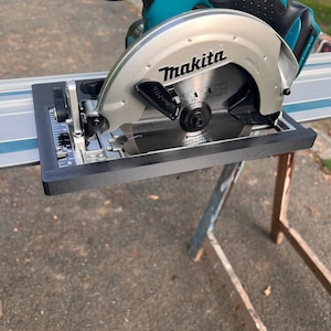 Könnte beinhalten: Eine türkis-schwarze Makita Kreissäge mit silberner Klinge. Die Säge ist auf einer schwarzen Basis montiert und trägt die Logos Makita und LXT. Die Säge ist zum Schneiden von Holz und anderen Materialien bestimmt.