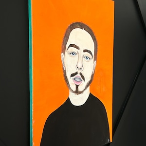 May include: An orange canvas painting of a man with brown hair, a beard, and blue eyes. The man is wearing a black shirt. The painting is displayed on a black wall.