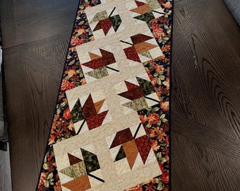 May include: A quilted table runner featuring a repeating pattern of autumn leaves in shades of brown, green, and orange. The leaves are set against a cream-colored background, bordered by a floral fabric with a dark navy blue edge.