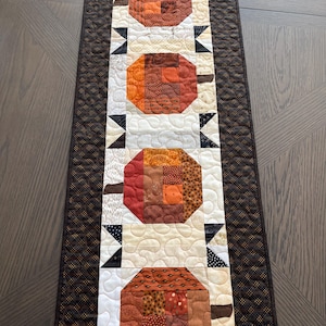 May include: A quilted table runner featuring a repeating pattern of stylized pumpkins. The pumpkins are made from various orange, brown, and tan fabrics. Black and white triangles separate the pumpkins. The runner has a brown border.