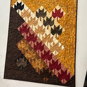 Puede incluir: Un edredón de pared rectangular con un patrón diagonal de bloques de hojas de arce en tonos burdeos, marrón oscuro, bronceado y crema. El fondo consiste en tela texturizada de color amarillo dorado y marrón oscuro con acolchado.