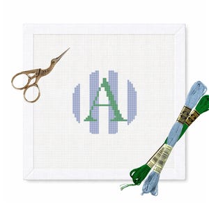 May include: A white needlepoint canvas with a blue and green monogram, gold scissors, and two skeins of embroidery floss. The monogram is a letter "A" in a striped design. The floss is blue and green.