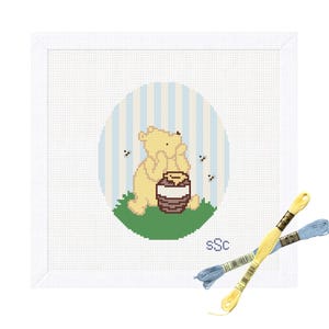 May include: A square white cross-stitch fabric featuring a central oval design of a yellow bear sitting on green grass with a brown honey pot, surrounded by light blue vertical stripes and small black bees, with two skeins of embroidery floss.