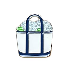 May include: A white tote bag pin with blue accents and a blue handle. The bag is filled with blue flowers and green leaves. The pin is made of metal and has a glossy finish.