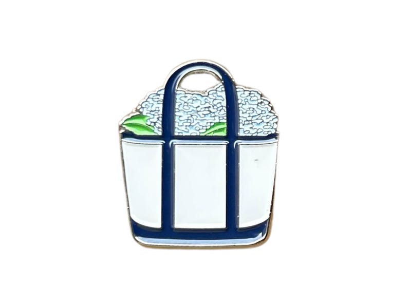 May include: A white tote bag pin with blue accents and a blue handle. The bag is filled with blue flowers and green leaves. The pin is made of metal and has a glossy finish.