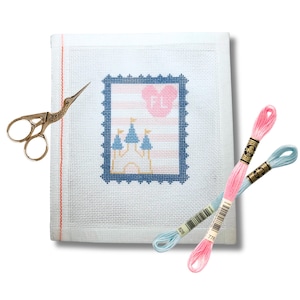 May include: A white needlepoint canvas with a blue and pink castle design, resembling a postage stamp. Gold scissors and two spools of embroidery floss in pink and blue are also present. The canvas has a red border.