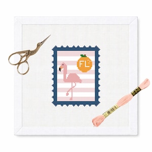 May include: A cross-stitch design featuring a pink flamingo on a striped background within a blue postage stamp frame. The stamp includes an orange circle with "FL" text. Gold scissors and a spool of pink thread are also present.