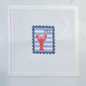 May include: A needlepoint canvas featuring a red lobster design within a blue and white striped stamp. The word "ME" is stitched above the lobster. The canvas is white with a red border.