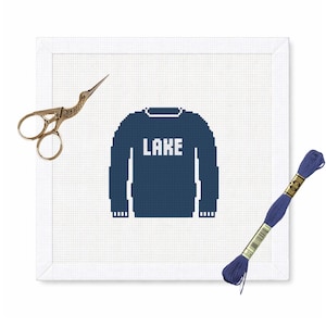May include: A cross-stitch design featuring a navy blue sweater with the word "LAKE" in white. The design is on a white canvas, with gold scissors and a spool of blue thread. The overall theme is a craft project.