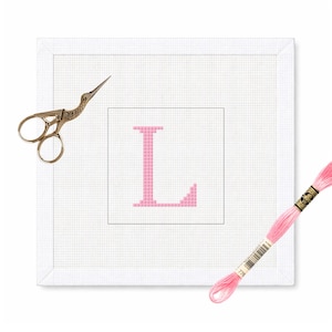 Peut inclure: Un projet de point de croix encadré blanc avec la lettre "L" rose au centre. Des ciseaux dorés et une bobine de fil à broder rose entourent l'œuvre, suggérant un thème artisanal.