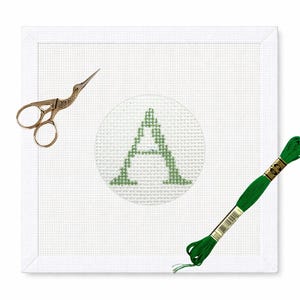 May include: A white needlepoint canvas with a green letter "A" stitched in the center. Gold-colored embroidery scissors and a skein of green embroidery floss are placed on the canvas, ready for crafting. The canvas is square and framed.