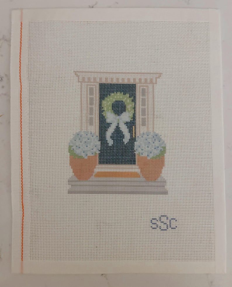 May include: Needlepoint canvas featuring a front door scene. The door is dark blue with a green wreath and white bow. Two potted blue hydrangeas flank the door. The canvas has an orange border on the left side and the initials "sSc" in the lower right corner.