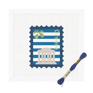 May include: A white cross-stitch fabric square featuring a stamp-shaped design with blue and white horizontal stripes, a beige Greek temple, green olive branches, and the word Greece in yellow. A single skein of dark blue embroidery floss is included.