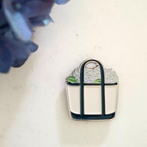 May include: A white enamel tote bag pin with black trim and handles, filled with a light gray floral design. The pin has a silver-colored border. A blue flower is in the upper left corner.