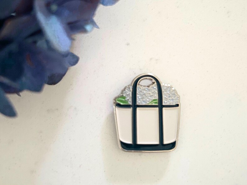 May include: A white enamel tote bag pin with black trim and handles, filled with a light gray floral design. The pin has a silver-colored border. A blue flower is in the upper left corner.