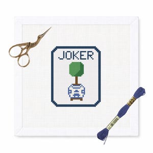 May include: A cross-stitch piece with the word "JOKER" in a navy blue frame, featuring a green topiary in a blue and white vase. Gold scissors and a skein of blue embroidery floss are placed on the white background.