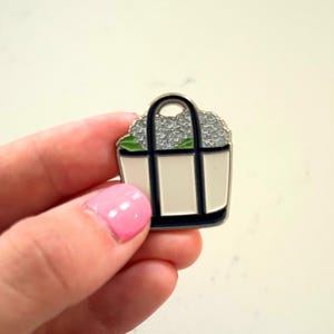 May include: A small, enamel pin in the shape of a tote bag. The bag is white with navy blue trim and a black handle. It is filled with gray flowers and green leaves.