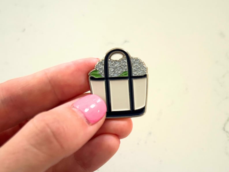 May include: A small, enamel pin in the shape of a tote bag. The bag is white with navy blue trim and a black handle. It is filled with gray flowers and green leaves.