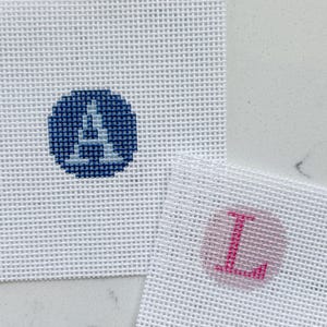 May include: Two white needlepoint canvases with embroidered letters. One canvas features a blue circle with a white "A" stitched inside. The other canvas has a pink circle with a pink "L" stitched inside. The canvases are on a white surface.