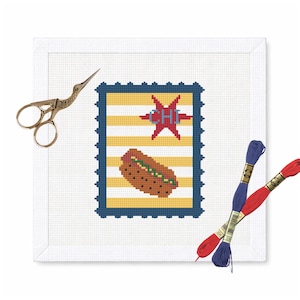 May include: A cross-stitch design featuring a hot dog, star, and the text "CHI" on a yellow and white striped background, framed in blue. Includes gold scissors and red and blue embroidery floss.