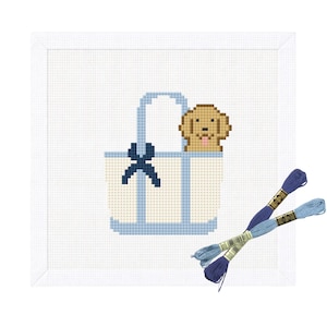 Könnte beinhalten: Eine Gobelin-Leinwand mit einem pixeligen Golden Retriever-Welpen, der aus einer hellblauen und cremefarbenen Tragetasche mit einer dunkelblauen Schleife herausschaut. Zwei Stränge blauen Stickgarns sind enthalten.