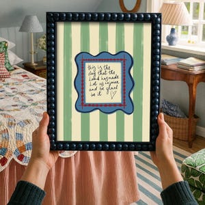 May include: A framed wall art print featuring a light green and cream vertical striped background. In the center, a wavy-edged blue frame contains the handwritten text: This is the day that the Lord has made. Let us rejoice and be glad in it.