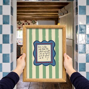May include: A framed wall art print featuring vertical sage green and cream stripes. In the center, a blue scalloped frame with a red dotted border surrounds a cream rectangle containing the handwritten text: This is the day that the Lord has made. Let us rejoice and be glad in it, followed by a small heart symbol.