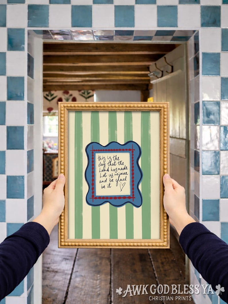May include: A framed wall art print featuring vertical sage green and cream stripes. In the center, a blue scalloped frame with a red dotted border surrounds a cream rectangle containing the handwritten text: This is the day that the Lord has made. Let us rejoice and be glad in it, followed by a small heart symbol.