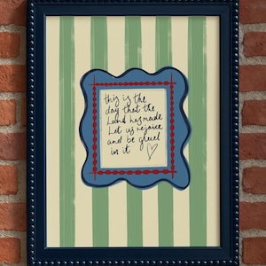 May include: A framed wall art print featuring vertical sage green and cream stripes. Centered is a wavy-edged blue plaque with a red dotted border containing the handwritten text: This is the day that the Lord has made. Let us rejoice and be glad in it.