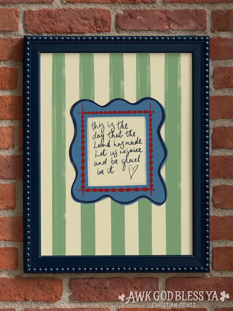 May include: A framed wall art print featuring vertical sage green and cream stripes. Centered is a wavy-edged blue plaque with a red dotted border containing the handwritten text: This is the day that the Lord has made. Let us rejoice and be glad in it.