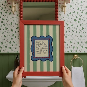 May include: A rectangular red picture frame containing a print with vertical green and cream stripes. The center features a blue scalloped border surrounding handwritten text that reads: This is the day that the Lord has made. Let us rejoice and be glad in it.