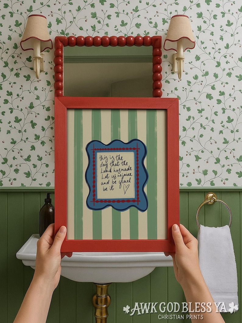 May include: A rectangular red picture frame containing a print with vertical green and cream stripes. The center features a blue scalloped border surrounding handwritten text that reads: This is the day that the Lord has made. Let us rejoice and be glad in it.