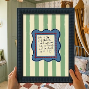 May include: A framed wall art print featuring a light green and cream vertical striped background. In the center, a blue wavy-edged frame surrounds a cream-colored square with the handwritten text: This is the day that the Lord has made.