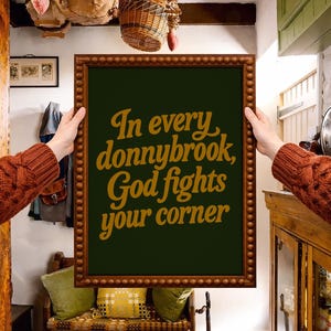 Peut inclure: Une affiche encadrée avec le texte "In every donnybrook, God fights your corner" en lettres dorées sur fond vert foncé. Le cadre est marron avec un bord perlé. L'affiche est tenue par une personne portant un pull marron.