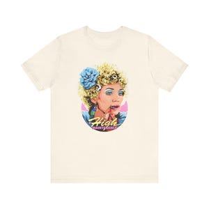 May include: A white t-shirt with a graphic of a woman with blonde hair and blue eyeshadow applying lipstick. The graphic is in a circle with the text "High Maintenance" below it.