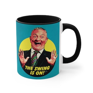 Op de afbeelding: Witte keramische mok met een zwarte rand en handvat. De mok heeft een kleurrijke illustratie van een man met een verraste uitdrukking, met de tekst "THE SWING IS ON!" eronder.