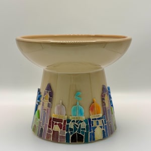 Pode incluir: Uma tigela de pedestal de cerâmica bege com um topo largo e raso. A base apresenta um design de mosaico de edifícios e cúpulas coloridas. A tigela é adequada para fins decorativos ou para servir.