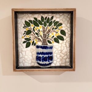 May include: A square wall art piece featuring a mosaic of a tree with green leaves and yellow lemons in a blue and white patterned pot. The background is made of white mosaic tiles, all encased in a simple brown wooden frame.