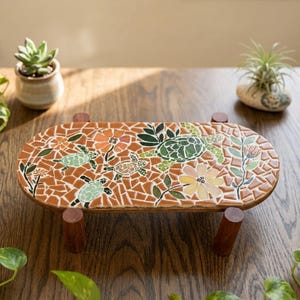 May include: An oval-shaped wooden stand featuring a mosaic tile top with a design of sea turtles, flowers, and leaves. The mosaic uses shades of orange, green, and yellow tiles. The stand is supported by four short, cylindrical wooden legs.
