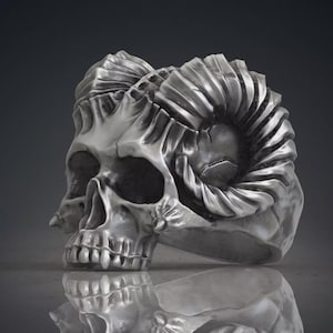 Può includere: Un anello color argento con un design dettagliato di teschio umano e grandi corna di ariete ricurve che si estendono dai lati. La superficie metallica ha una finitura lucida con aree scure incassate che mettono in risalto le trame dell'osso.