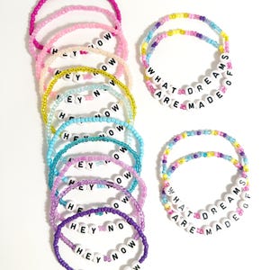 May include: A collection of colorful beaded bracelets. Some bracelets spell out "HEY NOW" and others say "WHAT DREAMS ARE MADE OF" in black letters on white beads. The bracelets are in shades of pink, blue, yellow, and purple.