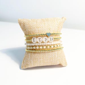 May include: Stack of beaded bracelets with gold accents and the letters "TTPD" spelled out on the bracelets. A small blue heart charm is on one bracelet. The bracelets are displayed on a small, tan burlap pillow.