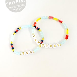 May include: Two beaded bracelets featuring a mix of light blue, red, yellow, and dark blue beads. One bracelet displays the white letter beads spelling TAY STORY, while the other features the white letter beads spelling TS 5 with gold lettering.