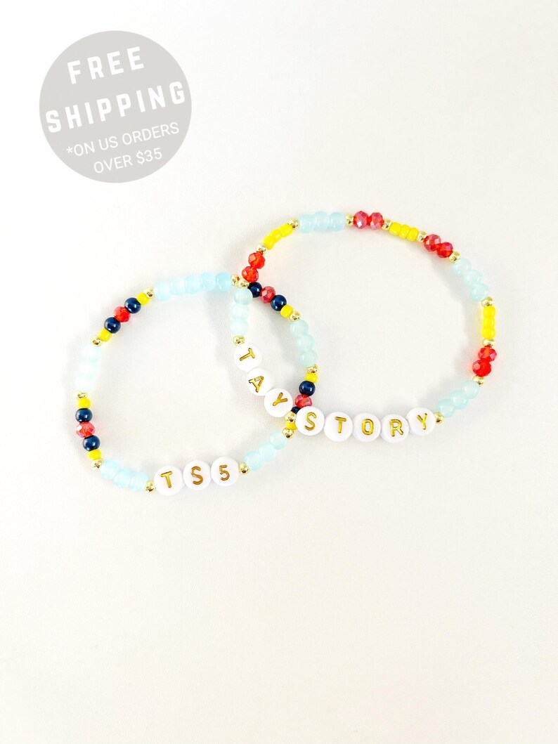 May include: Two beaded bracelets featuring a mix of light blue, red, yellow, and dark blue beads. One bracelet displays the white letter beads spelling TAY STORY, while the other features the white letter beads spelling TS 5 with gold lettering.