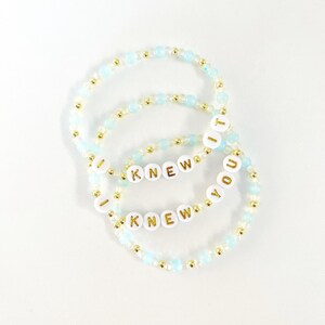 May include: Two beaded bracelets featuring light blue and metallic gold beads. Each bracelet includes white circular beads with gold lettering that spells out the phrase I KNEW YOU, with the word IT appearing on one of the bracelets.
