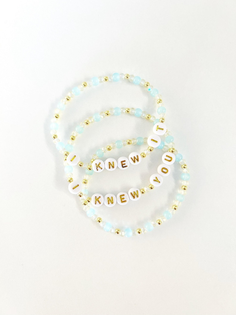 May include: Two beaded bracelets featuring light blue and metallic gold beads. Each bracelet includes white circular beads with gold lettering that spells out the phrase I KNEW YOU, with the word IT appearing on one of the bracelets.